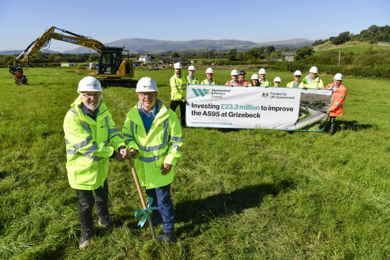 A group of 15 key stakeholders involved in the Grizebeck A595 project holding an investment sign in front of a digger in a field where a new road will be built