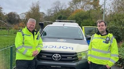 Two CCTV operators in hi-vis yellow jackets standing beside a police car in a park. CCTV camera in the background.