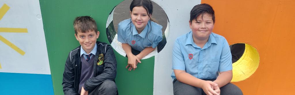 Three children in front of a football wall, a brightly coloured wall with holes for kicking a football through..