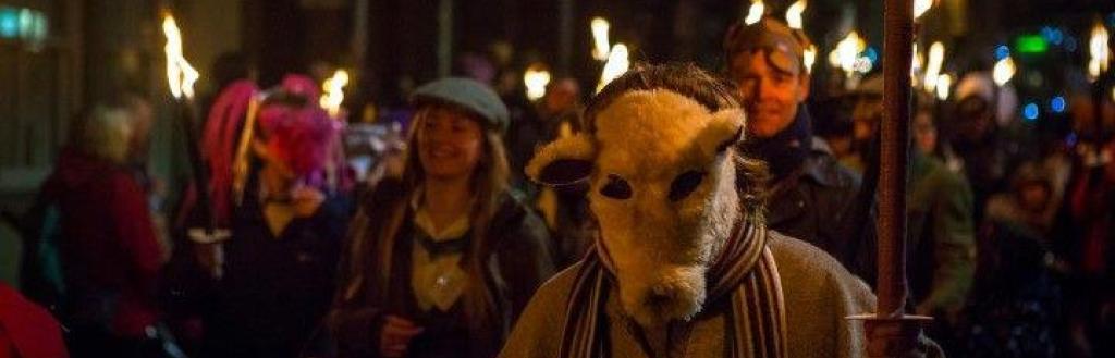 A line of people carrying lit torches at night as part of a parade, one of them wearing an animal mask.