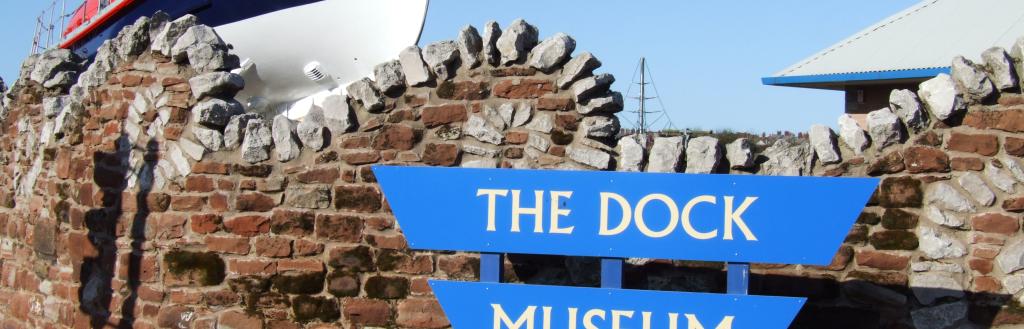 A curved stone wall. A boat. A sign that says The Dock Museum.